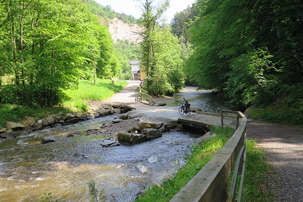 Blick auf einen Fluss - die Feistritz, daneben geht der Radweg entlang. Der Radfahrer fährt gerade über die Brücke.