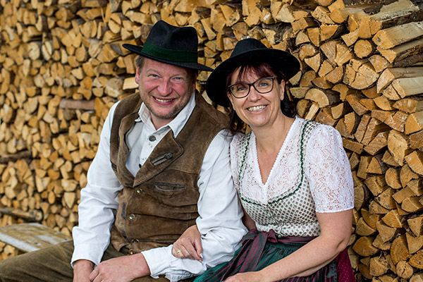 Margret und Wolfgang in Tracht sitzen auf einer schmalen Holzbank vor dem Holzstoß