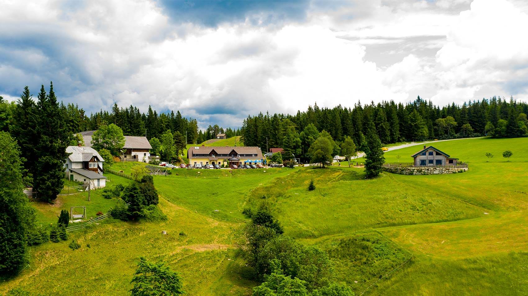 Außenansicht des Gasthofs - fotografiert von der gegenüberliegenden Seite, man sieht alle Gebäude mit den Wiesen und Wäldern