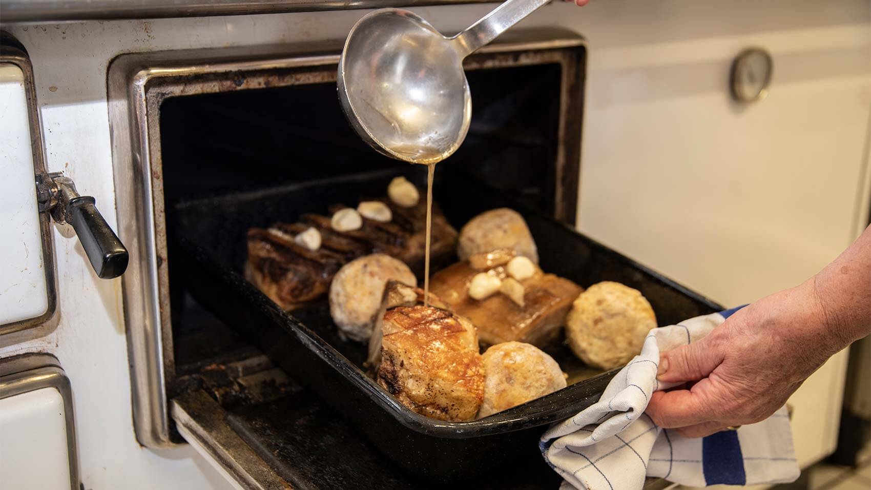 Ein Schweinsbraten wird gerade aus dem Holzofen gezogen und mit einem Schöpfer mit Flüssigkeit übergossen.