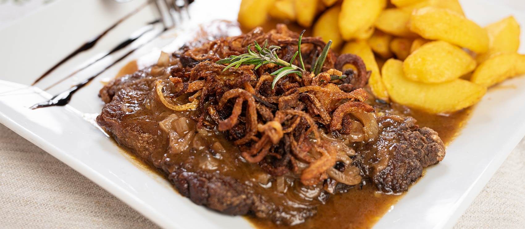 Zwiebelrostbraten mit Bratkartoffel auf einen rechteckigen Teller serviert.