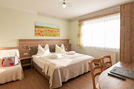 Ein Zimmer mit Doppelbett. Rechts befindet sich ein Tisch mit zwei Sesseln. An der Wand hängt ein Bild mit roten Blumen.