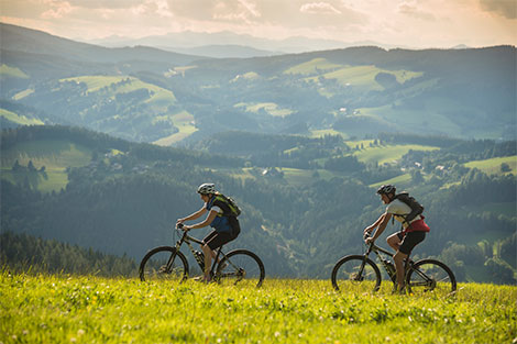 Zwei Mountainbiker fahren über eine Wiese. Im Hintergrund sind Berge und Täler zu sehen.