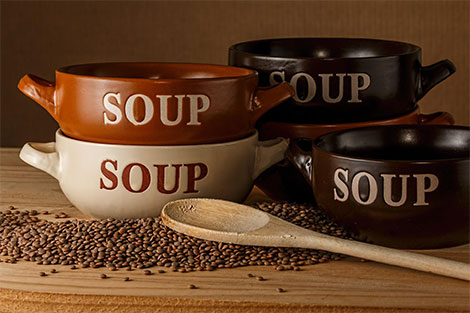Suppenschüsseln aufeinander gestellt - mit dem Schriftzug Soup und davor liegt ein Kochlöffel und Pfefferkörner.