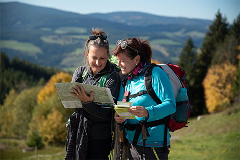 Zwei Frauen in Wandermontur und mit Ausrüstung sehen sich eine Wanderkarte an. Im Hintergrund sie grüne Wälder zu sehen.