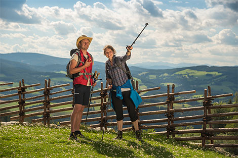Zwei Wanderer stehen bei einem Jägerzaun und blicken in die Landschaft, dahinter sieht man die Hügel der Oststeiermark.