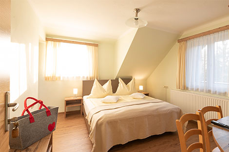 Schlafzimmer mit zwei Fenstern und Doppelbett. Auf einem Regal steht eine grau-rote Tasche.